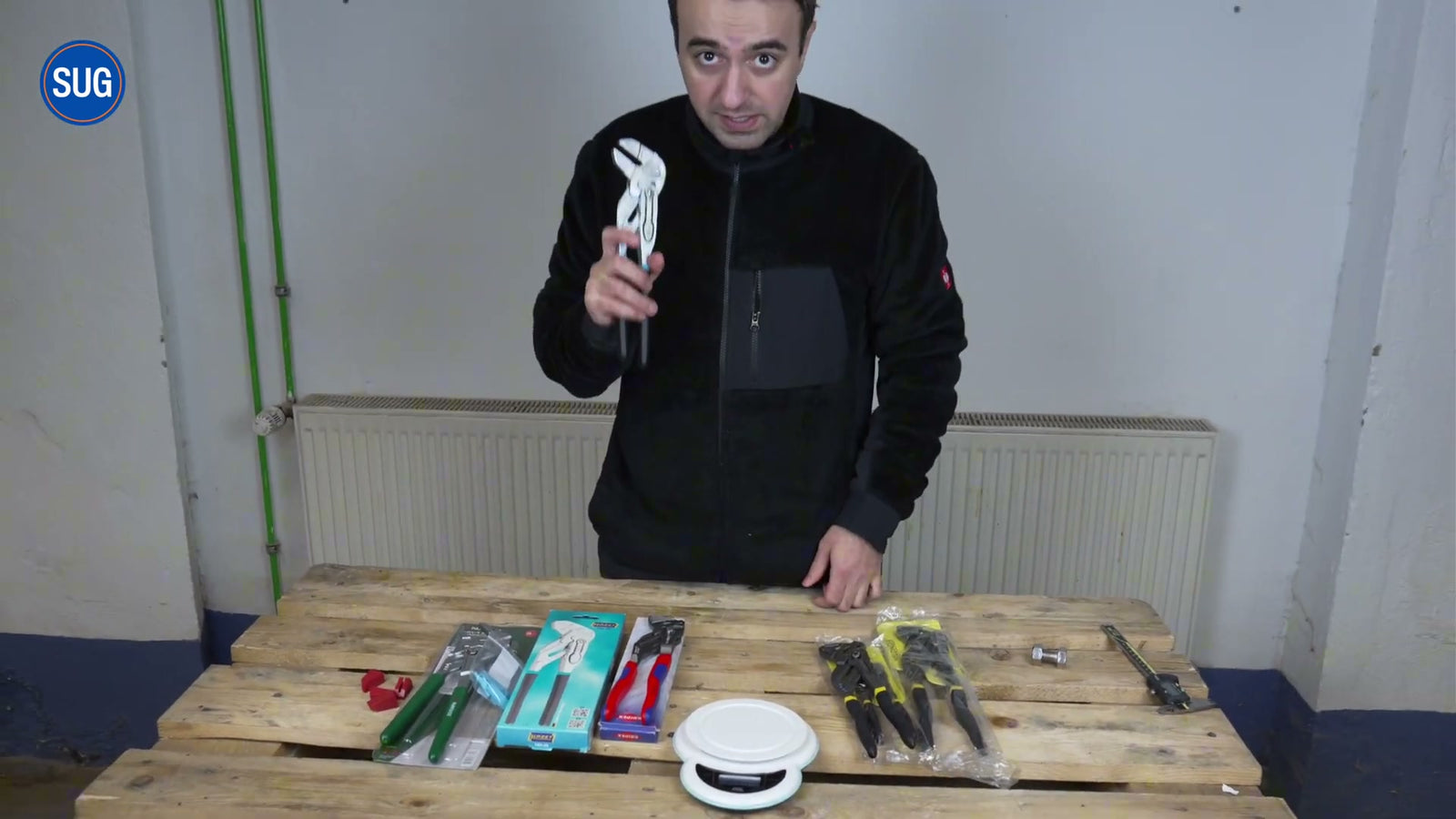 Der Tester hält einen Knipex-Zangenschlüssel hoch; auf der Palette liegen die Verpackungen der vier getesteten Zangen (u. a. Weldinger-Set mit grünen Griffen, Hazet-Karton, Knipex-Blister, Parkside) und eine Küchenwaage.