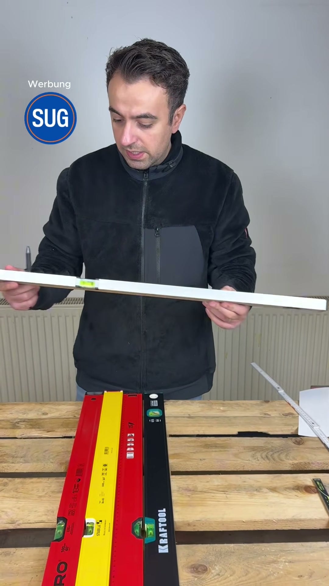 Der Tester hält die graue No-Name-Wasserwaage zur Libellen-Prüfung hoch; davor auf der Palette liegen die rote PRO, die gelbe Stabila und die schwarze Kraftool mit sichtbaren Libellen.