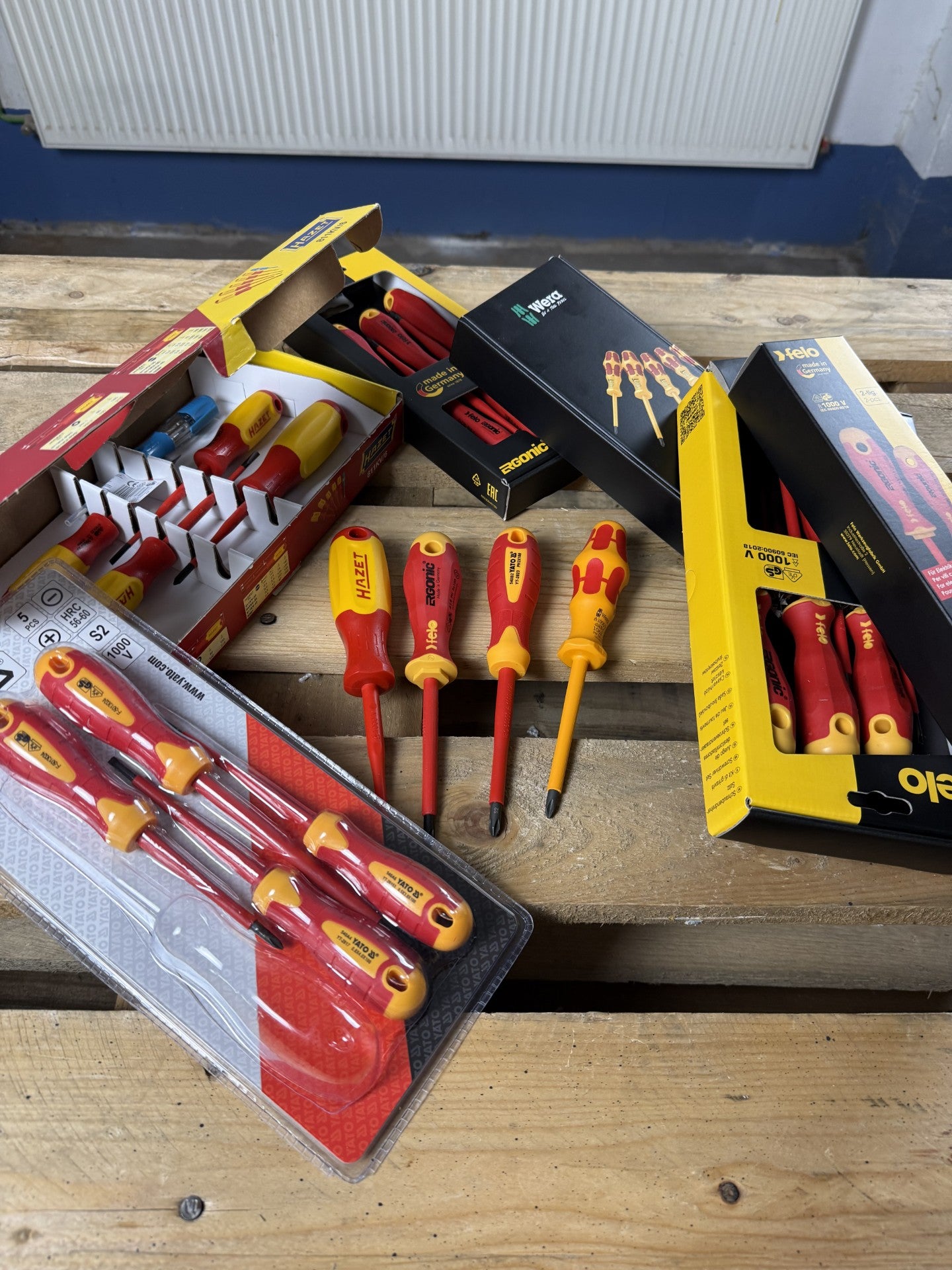 Alle vier VDE-Schraubendreher-Sets mit ihren Verpackungen auf der Werkbank: geöffnete Hazet-Box, Wera- und Felo-Verpackungen, Yato-Blister — davor vier Schraubendreher nebeneinander