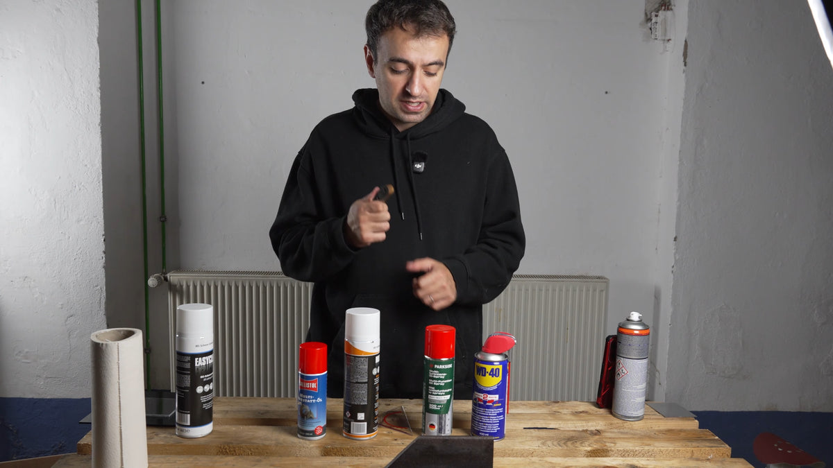 Holzwerkbank mit den vier Multisprays Ballistol, IBS MultiFix, Parkside und WD-40 in einer Reihe, daneben weitere Dosen und eine Papprolle, dahinter der Tester im schwarzen Hoodie